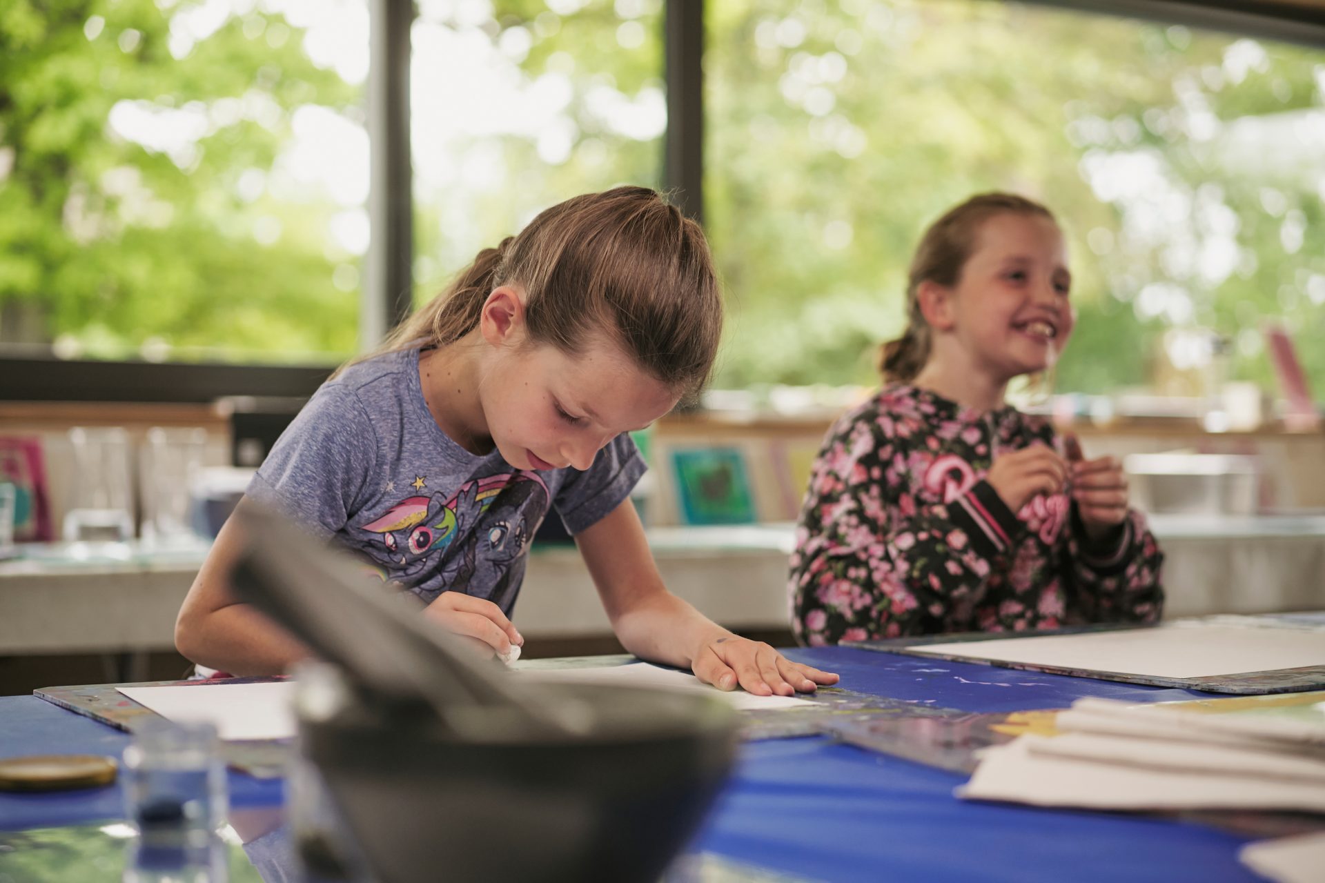 Zwei junge Mädchen sitzen an einem Tisch in einem hellen Raum. Die eine zeichnet konzentriert auf dem Papier, während die andere lächelt und aufschaut. Auf dem Tisch liegen Malutensilien verstreut, und durch die großen Fenster sieht man draußen Bäume.