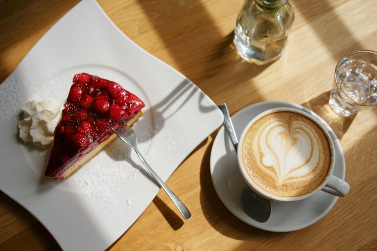 Ein Stück Käsekuchen mit Beeren und Schlagsahne auf einem Teller, daneben eine Tasse Cappuccino mit Latte Art, ein Glas Wasser und eine kleine Flasche auf einem Holztisch im Sonnenlicht.