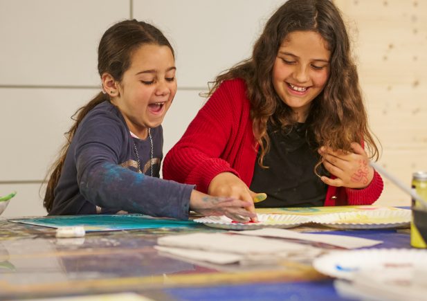 Zwei junge Mädchen sitzen an einem mit Malutensilien gedeckten Tisch und malen lächelnd und lachend zusammen. Ein Mädchen greift begeistert zum Pinsel, während das andere sich an der kreativen Tätigkeit erfreut.
