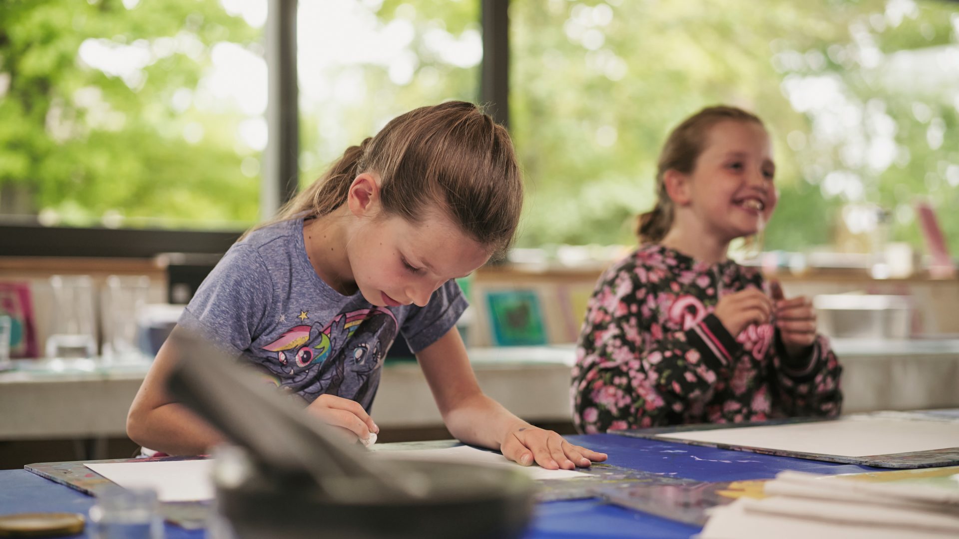 Zwei junge Mädchen sitzen an einem Tisch in einem hellen Raum. Die eine zeichnet konzentriert auf dem Papier, während die andere lächelt und aufschaut. Auf dem Tisch liegen Malutensilien verstreut, und durch die großen Fenster sieht man draußen Bäume.