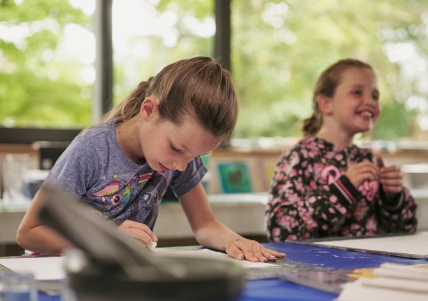 Zwei junge Mädchen sitzen an einem Tisch in einem hellen Raum. Die eine zeichnet konzentriert auf dem Papier, während die andere lächelt und aufschaut. Auf dem Tisch liegen Malutensilien verstreut, und durch die großen Fenster sieht man draußen Bäume.