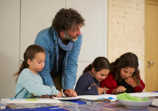 Ein Mann beugt sich über einen Tisch und hilft drei jungen Mädchen bei einem Kunstprojekt, während sie in einem hellen, modernen Klassenzimmer auf Papier zeichnen und malen.