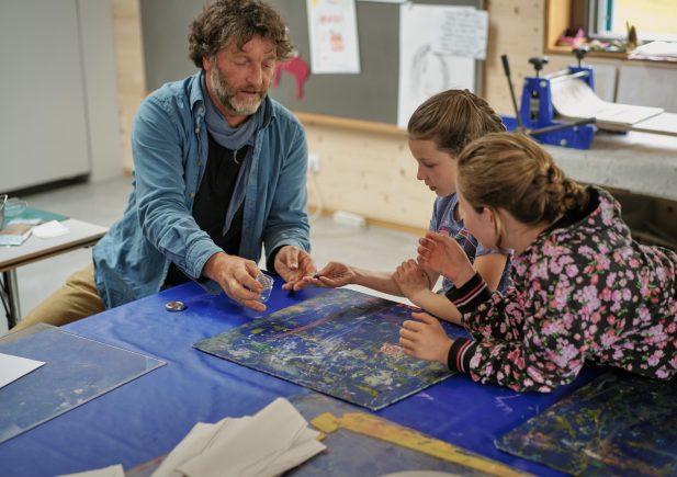 Ein Mann sitzt mit zwei Mädchen an einem Tisch und leitet sie an, während sie an einem Kunstprojekt mit bunten Materialien arbeiten, die in einem Klassenzimmer verteilt sind.