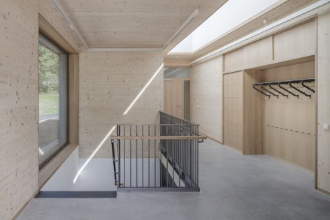 A modern, minimalist hallway with light wood walls and built-in storage, a metal railing around a staircase, a large window, and hooks for hanging items. Sunlight streams in, creating a bright and airy atmosphere.