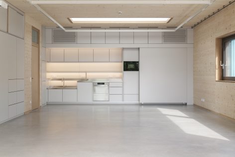 Minimalist, modern kitchen with light wood walls and ceiling, light grey cabinets, built-in appliances, and an empty open space with a large window on the right. The room has a clean, uncluttered look.