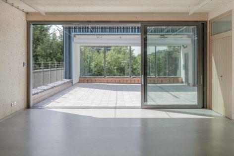 A spacious, modern room with large sliding glass doors opening to a sunlit patio, surrounded by metal railings and lush green trees outside. The interior features light wood walls and a smooth grey floor.