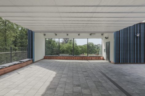 A covered outdoor area with a tiled floor, large windows overlooking trees and greenery, and blue-striped walls, allowing natural light to fill the open, spacious setting.