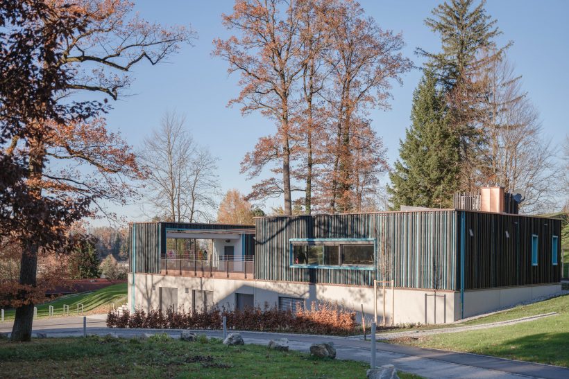 Ein modernes Gebäude mit Flachdach, vertikalen Holzlatten und großen Fenstern steht an einem sonnigen Tag mit blauem Himmel inmitten von hohen Herbstbäumen und Gras.
