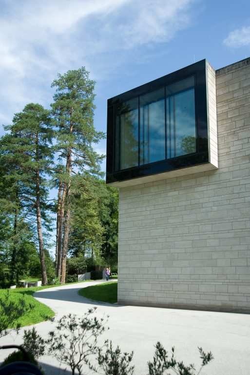 Ein modernes helles Steingebäude mit einem großen Glasfenster ragt nach außen. Ein geschwungener Weg, grüne Bäume und ein blauer Himmel umgeben das Gebiet, und in der Ferne gehen zwei Menschen.