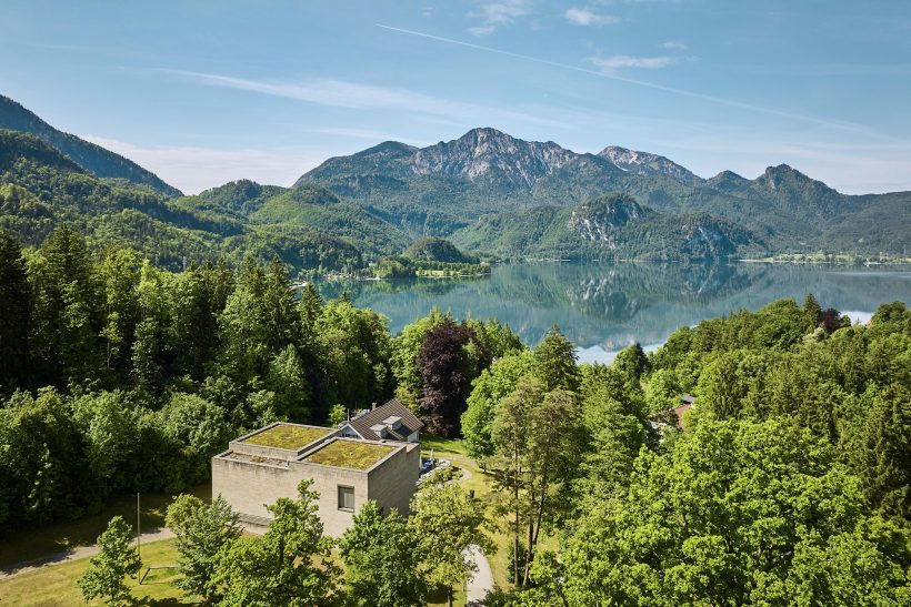 Ein modernes Betongebäude, umgeben von üppigen grünen Bäumen, überblickt einen ruhigen See mit Bergen im Hintergrund unter einem klaren blauen Himmel.