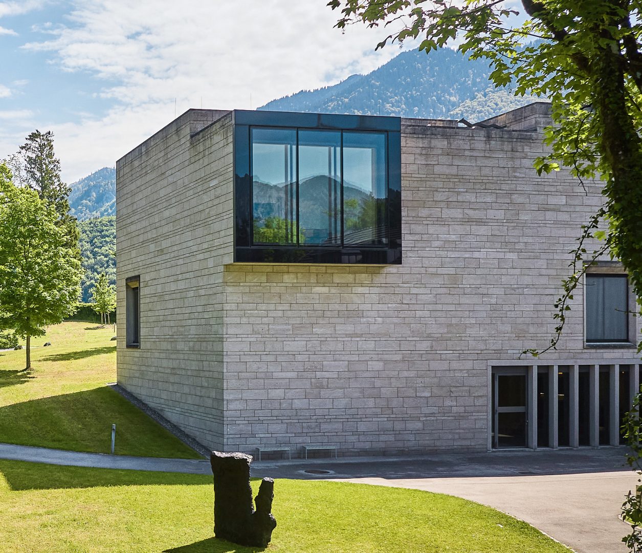 Ein modernes Steingebäude mit großen Glasfenstern steht auf einer Wiese, umgeben von Bäumen und Bergen unter einem teilweise bewölkten Himmel.
