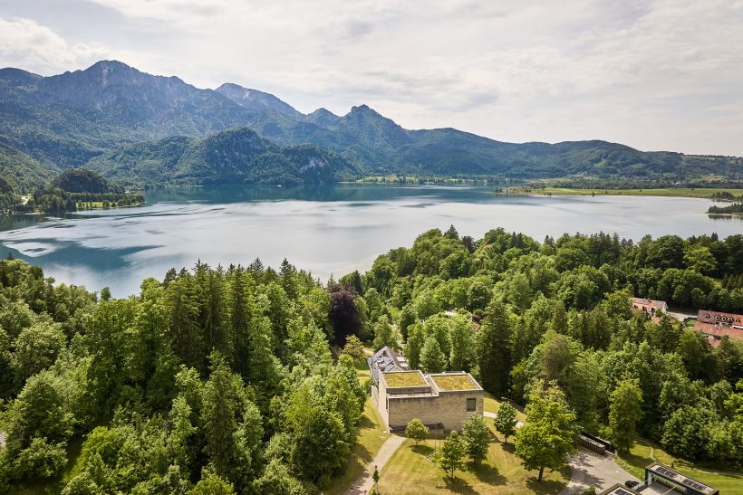 Ein modernes Betongebäude, umgeben von üppigen grünen Bäumen, überblickt einen ruhigen See mit Bergen im Hintergrund unter einem klaren blauen Himmel.