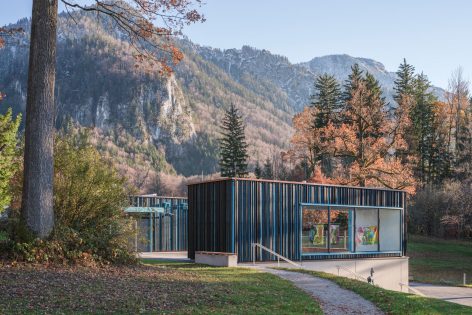 Ein modernes Gebäude mit Glaswänden und vertikalen blauen Paneelen steht inmitten von Herbstbäumen, in denen bunte Kunstwerke zu sehen sind. Im Hintergrund erheben sich Berge unter einem klaren Himmel.