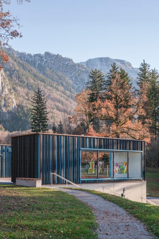 Ein modernes Gebäude mit Glaswänden und vertikalen blauen Paneelen steht inmitten von Herbstbäumen, in denen bunte Kunstwerke zu sehen sind. Im Hintergrund erheben sich Berge unter einem klaren Himmel.