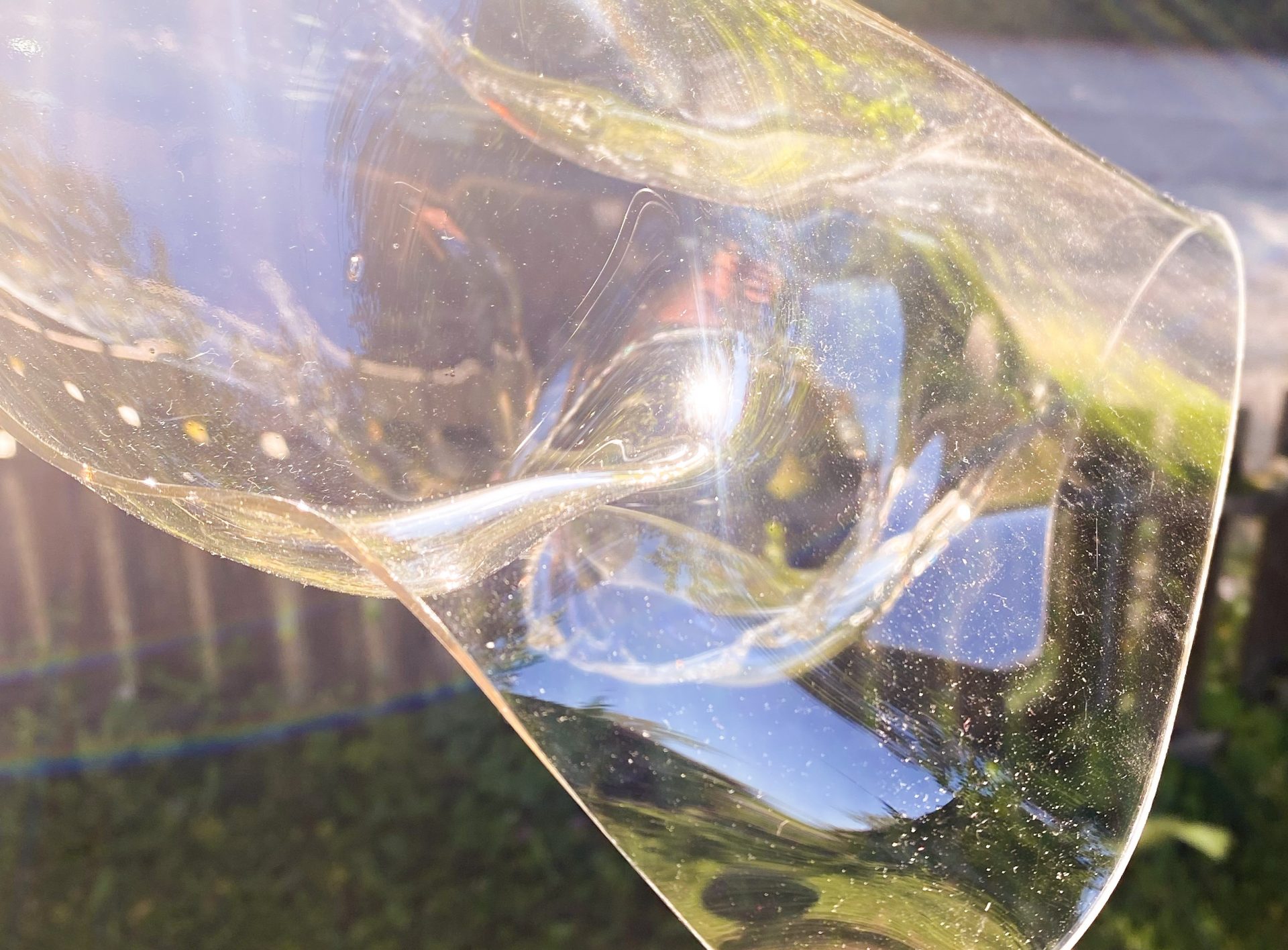 Nahaufnahme einer zerknitterten, durchscheinenden Plastikfolie, die das Sonnenlicht reflektiert, mit einem unscharfen Hintergrund aus einem Holzzaun, grünem Gras und einem gepflasterten Weg.