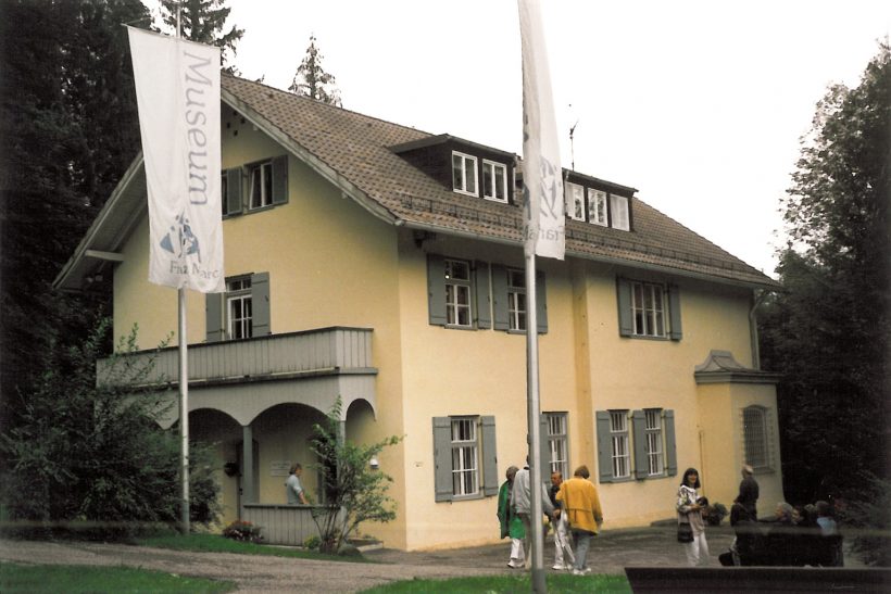 Eine Gruppe von Menschen steht vor einem gelben, zweistöckigen Haus mit Giebeldach, Balkon und weißer Verkleidung. Im Vordergrund sind zwei weiße Fahnen mit der Aufschrift Museum zu sehen. Das Haus ist von Bäumen umgeben.