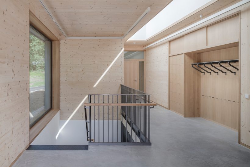 Ein heller, moderner Flur mit Holzwänden und -decken, einem großen Fenster, Betonboden, einer Treppe mit Metallgeländer und Schränken mit Kleiderhaken auf der rechten Seite. Das Sonnenlicht bildet eine diagonale Linie über die Szene.