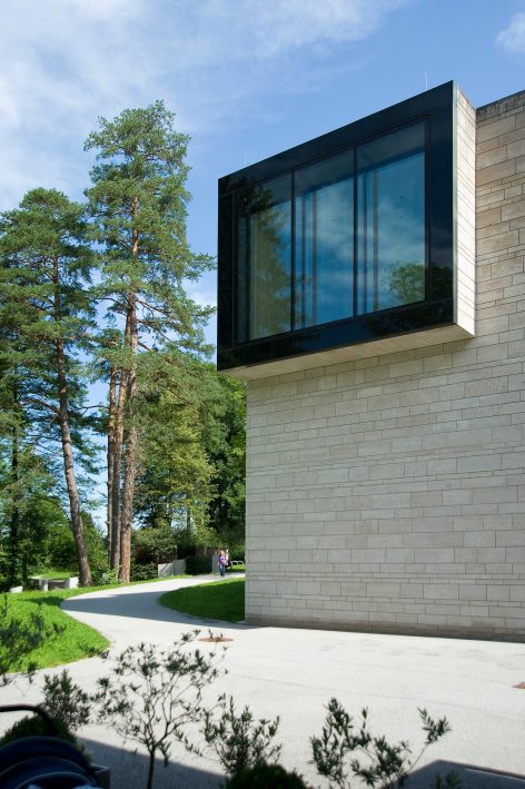 A modern building with light stone walls and a large, glass-walled overhanging room. Trees and greenery surround the structure under a blue sky, with a curved pathway in the foreground.