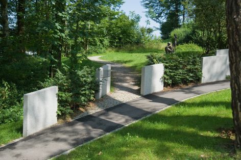 A paved path curves through a green park, bordered by white stone slabs and low bushes. In the distance, there is a sculpture surrounded by trees and grass under a clear blue sky.