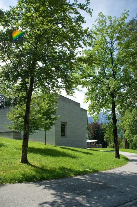 A modern, light gray building with a single window is partially visible behind lush green trees and grass, along a curving paved path on a sunny day. A small rainbow lens flare appears near the top left.
