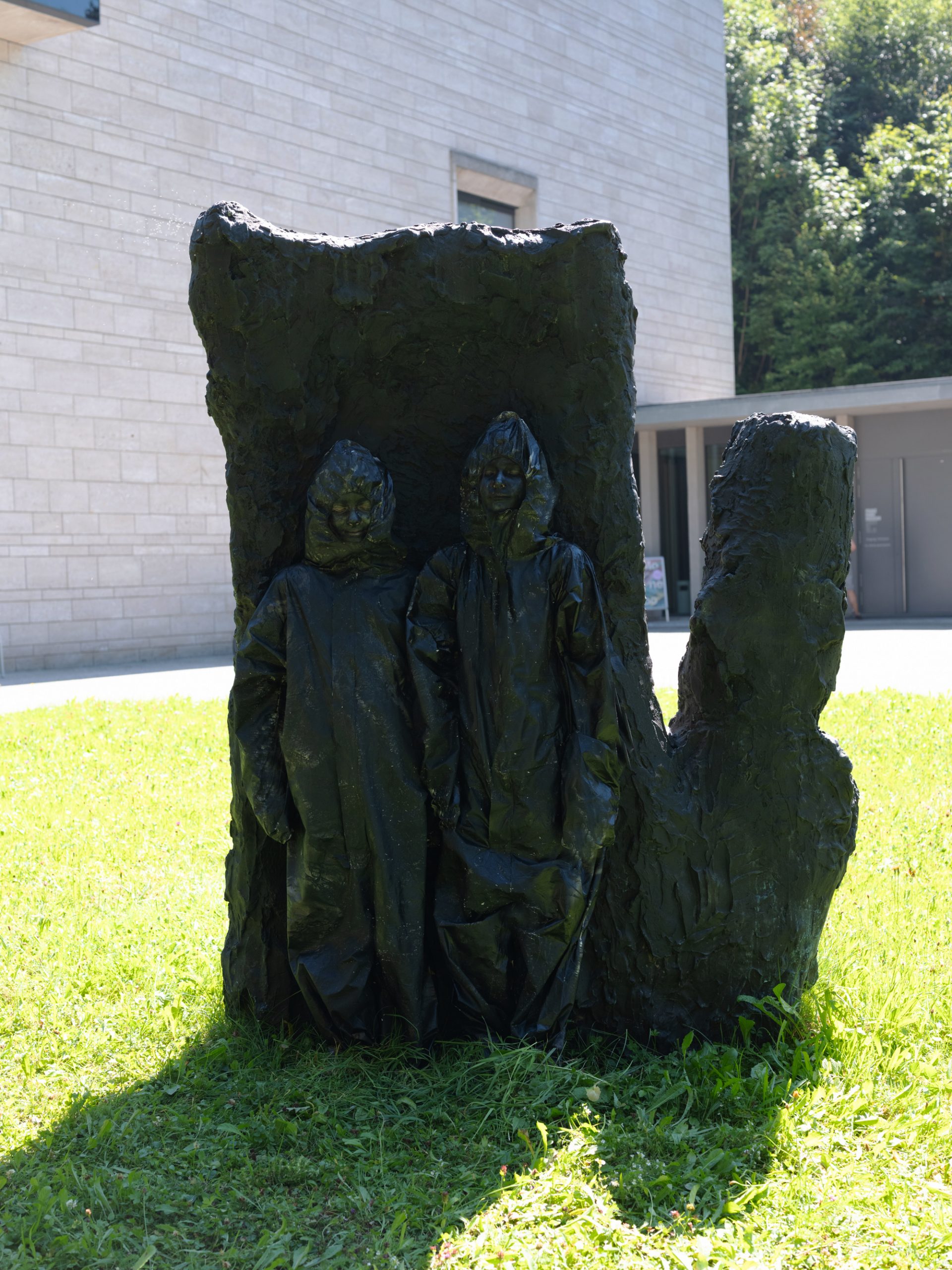 Zwei menschliche Figuren in strukturierten, dunklen Kapuzenmänteln stehen nebeneinander, teilweise umschlossen von einer groben, abstrakten Skulptur im Freien auf einer Wiese mit einem modernen Gebäude im Hintergrund.