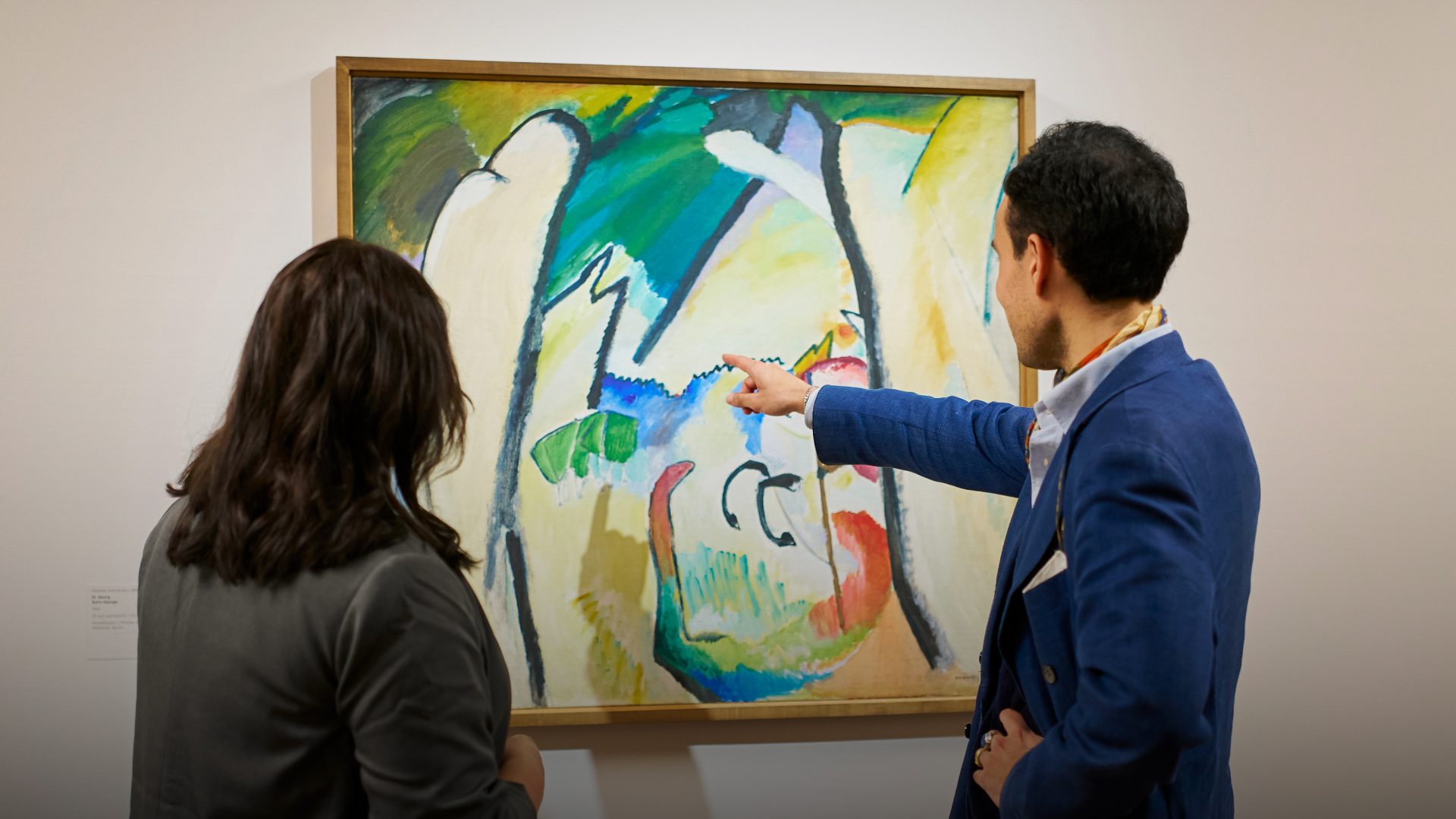 Zwei Personen stehen vor einem abstrakten Gemälde in einer Galerie; eine Person zeigt auf das Kunstwerk, während die andere es beobachtet, beide konzentriert auf die farbenfrohe Komposition.