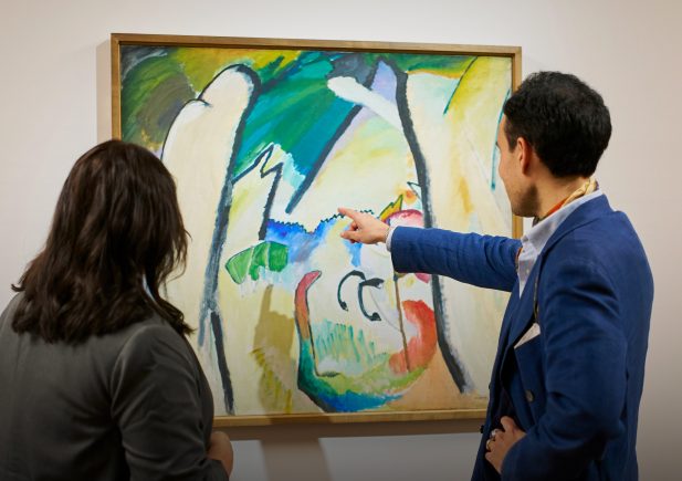 Zwei Personen stehen vor einem abstrakten Gemälde in einer Galerie; eine Person zeigt auf das Kunstwerk, während die andere es beobachtet, beide konzentriert auf die farbenfrohe Komposition.