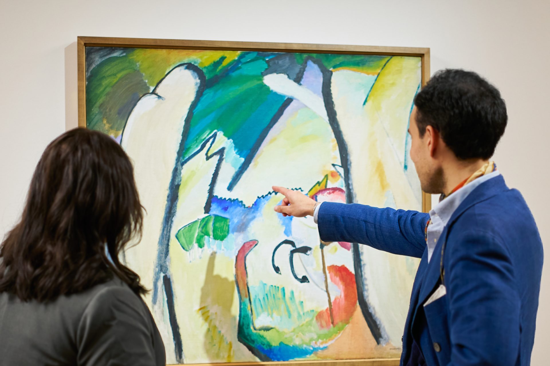 Zwei Personen stehen vor einem abstrakten Gemälde in einer Galerie; eine Person zeigt auf das Kunstwerk, während die andere es beobachtet, beide konzentriert auf die farbenfrohe Komposition.