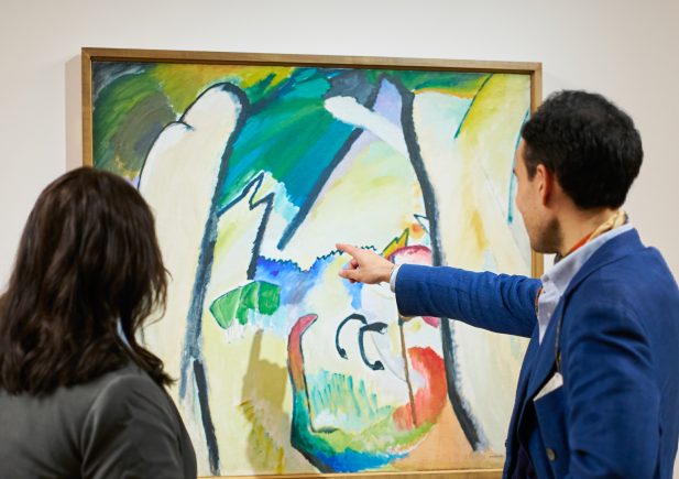 Zwei Personen stehen vor einem abstrakten Gemälde in einer Galerie; eine Person zeigt auf das Kunstwerk, während die andere es beobachtet, beide konzentriert auf die farbenfrohe Komposition.