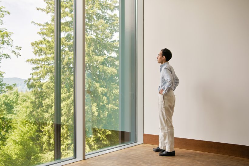 Eine Person steht in einem Haus, die Hände in die Hüften gestemmt, und blickt durch hohe, raumhohe Fenster auf grüne Bäume und eine natürliche Landschaft im Freien.