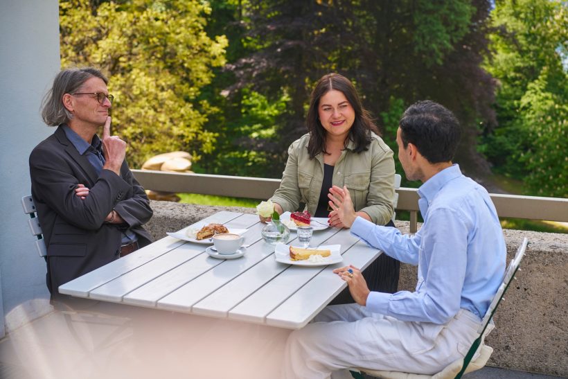 Drei Personen sitzen an einem weißen Tisch im Freien und unterhalten sich, während sie Teller mit Gebäck und Kaffee vor sich haben. Im Hintergrund sind grüne Bäume und Sonnenlicht zu sehen.