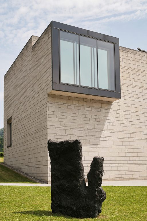 Ein modernes, beigefarbenes Backsteingebäude mit einem großen, verglasten Überhang. Im Vordergrund steht eine dunkle abstrakte Skulptur auf einem gepflegten grünen Rasen unter einem teilweise bewölkten Himmel.