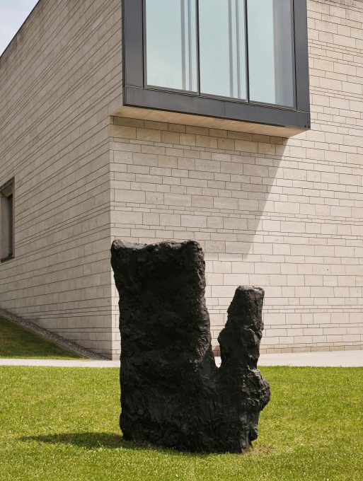 Eine dunkle, abstrakte Steinskulptur steht auf grünem Gras vor einem modernen Gebäude mit hellen Ziegelwänden und großen Fenstern.