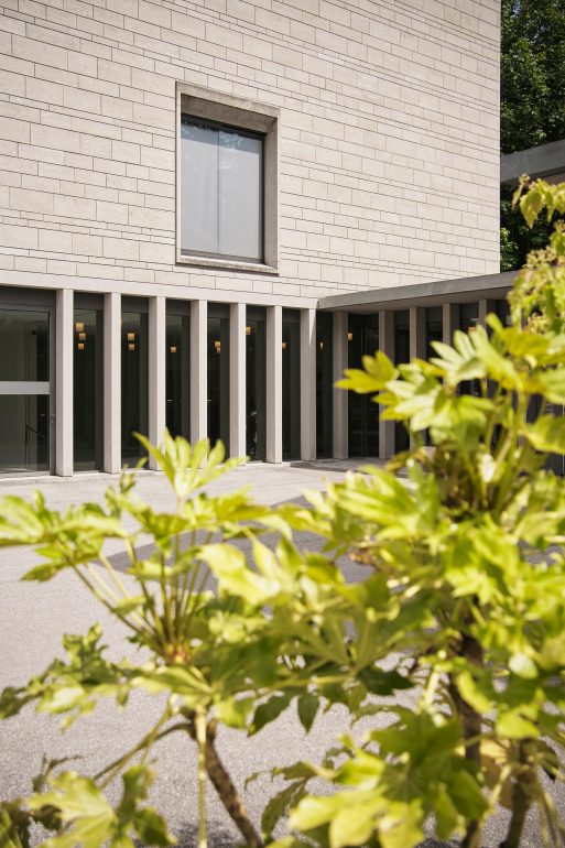 Ein modernes Gebäude mit hellen Steinwänden, hohen schmalen Fenstern und einem großen Fenster im Obergeschoss. Im Vordergrund befinden sich grüne Blattpflanzen, die die Sicht teilweise verdecken.