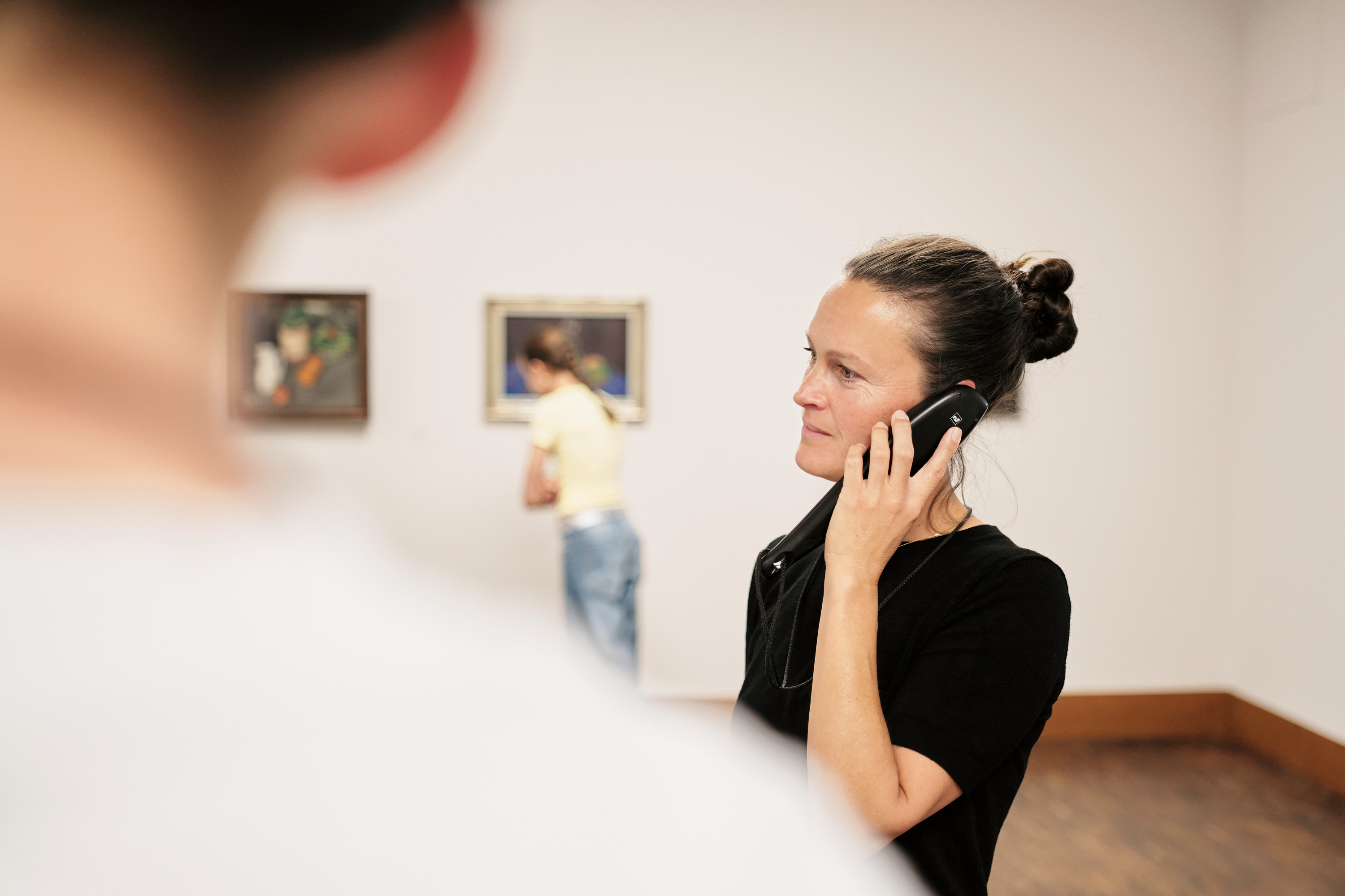 Eine Frau in einem schwarzen Hemd hört einem Audioguide in einer Kunstgalerie zu, während eine andere Person die Gemälde an der Wand im Hintergrund betrachtet. Das Foto wird teilweise von einer unscharfen Figur im Vordergrund verdeckt.