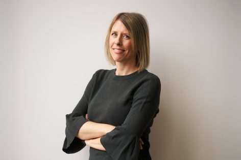 Eine Frau mit glattem blondem Haar, die ein schwarzes langärmeliges Oberteil trägt, steht mit verschränkten Armen vor einer schlichten weißen Wand, lächelt leicht und schaut in die Kamera.