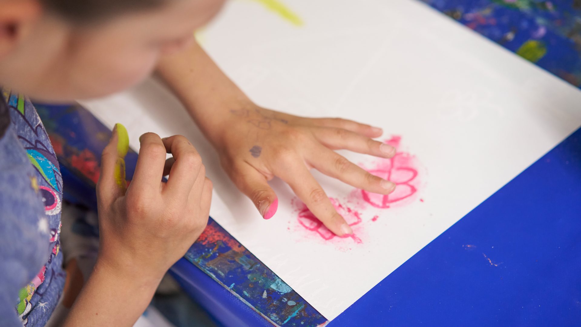 Ein Kind malt auf einem weißen Blatt Papier und zeichnet mit rosa Kreide oder Buntstift seine Hand nach. Seine Finger sind rosa verschmiert und es sitzt an einem blauen Tisch.