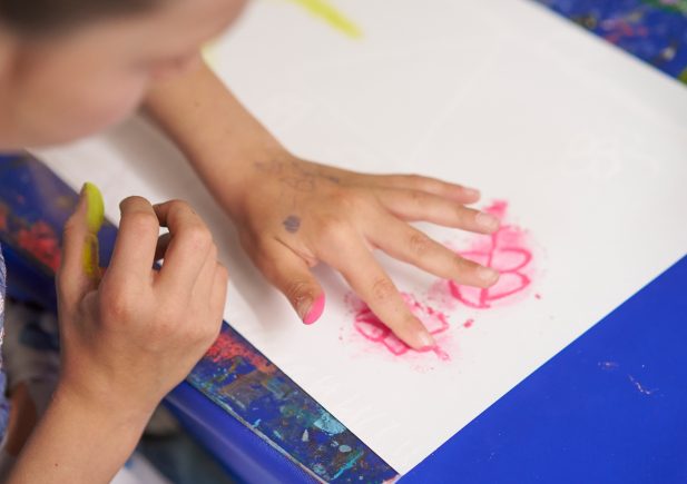 Ein Kind malt auf einem weißen Blatt Papier und zeichnet mit rosa Kreide oder Buntstift seine Hand nach. Seine Finger sind rosa verschmiert und es sitzt an einem blauen Tisch.