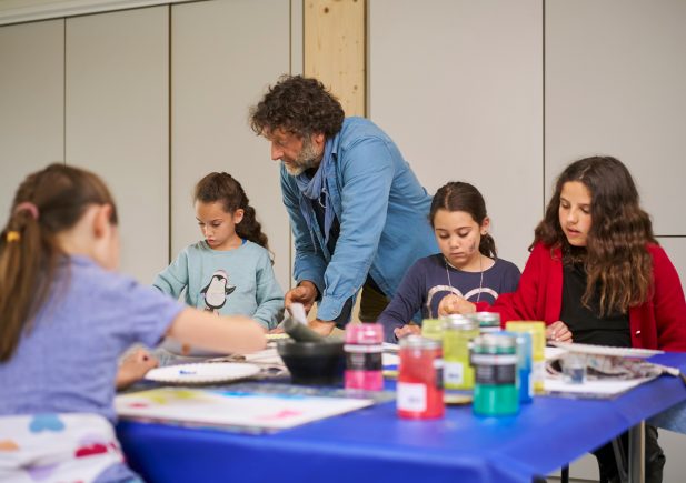 Ein Mann hilft vier jungen Mädchen beim Malen an einem Tisch, der mit Malutensilien und bunten Farbdosen bedeckt ist. Die Kinder konzentrieren sich auf ihre Kunstwerke in einem Atelierraum.