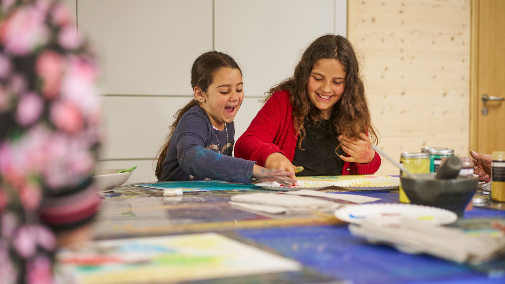 Zwei junge Mädchen sitzen an einem mit Malutensilien gedeckten Tisch und malen lächelnd und lachend zusammen. Ein Mädchen greift begeistert zum Pinsel, während das andere sich an der kreativen Tätigkeit erfreut.