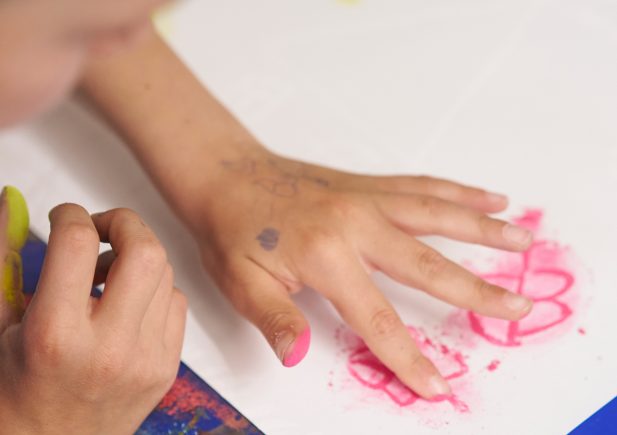 Ein Kind malt mit seinen Fingern rosa Kreide auf weißem Papier und kreiert so eine Blumenform. Die andere Hand ruht auf dem Tisch, und beide Hände haben Kreidestriche. Das Kunstwerk steht auf einem blauen Tisch.