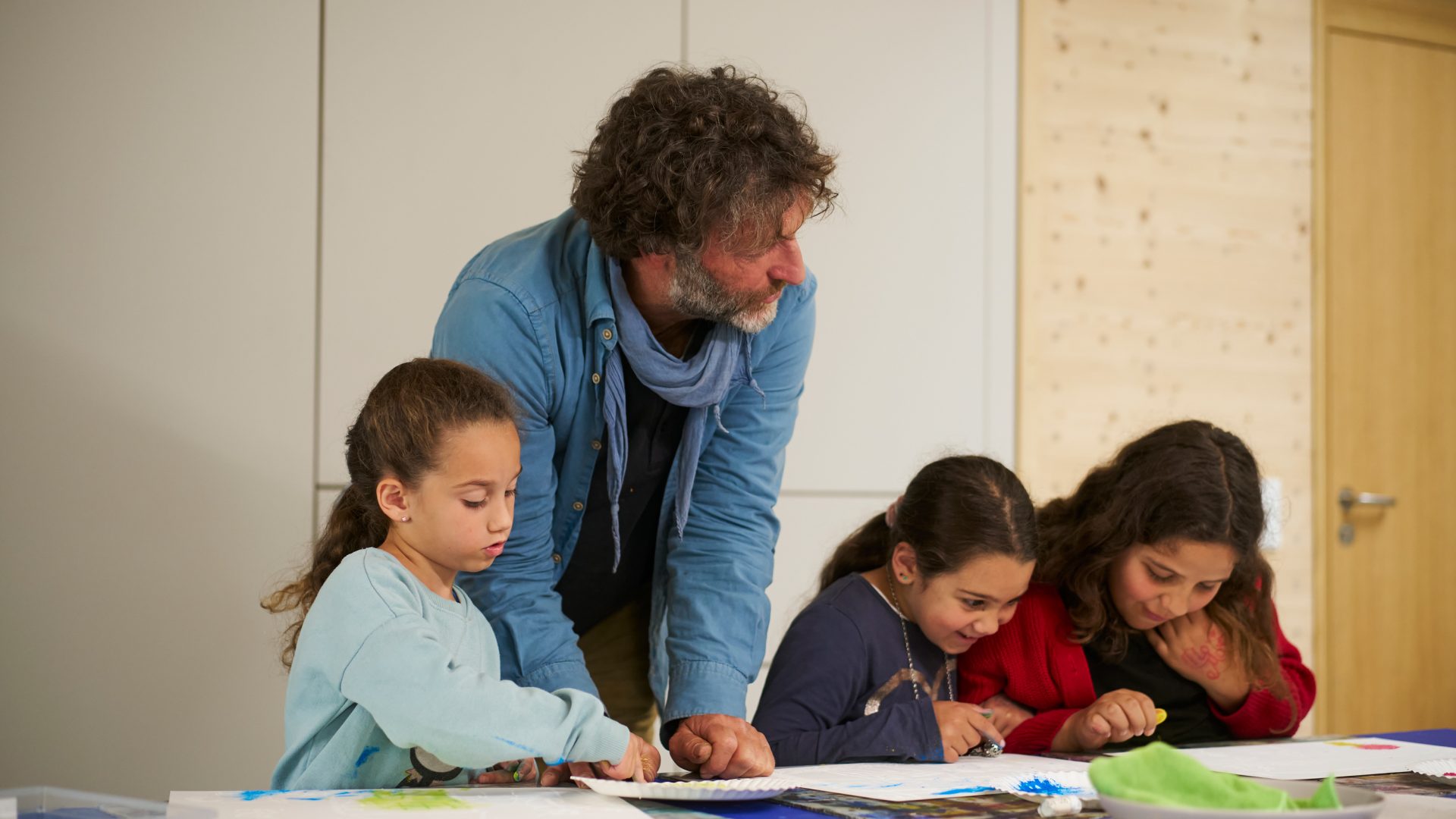 Ein Mann beugt sich über einen Tisch und hilft drei jungen Mädchen bei einem Kunstprojekt, während sie in einem hellen, modernen Klassenzimmer auf Papier zeichnen und malen.