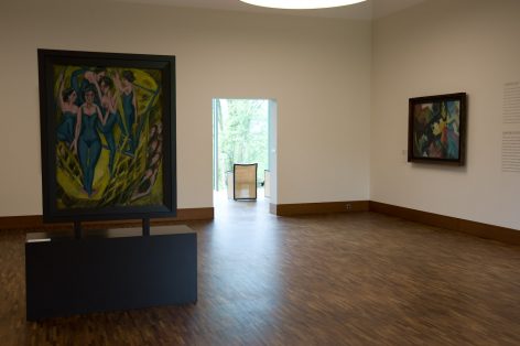 A modern art gallery with wooden floors displays two colorful paintings on white walls. One painting on a black stand features blue-toned figures; a window in the background reveals greenery outside.