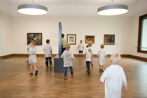 A group of children wearing paint-splattered smocks walk around an art gallery with framed paintings on white walls and wooden floors. Two large round lights hang from the ceiling.