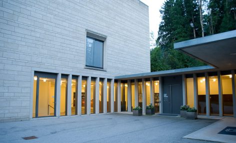 A modern building with light-colored brick walls, tall vertical windows, and illuminated interior. There are potted plants near a gray door, and trees can be seen in the background.