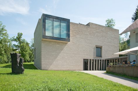 A modern, minimalist building with light stone walls and large glass windows stands on a green lawn. Trees and another building are nearby, and people are seated under umbrellas on a terrace.