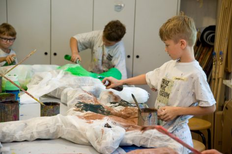 Three children paint and spray large, human-shaped figures covered in white material at a table in an art classroom. Paint cans and brushes are scattered on the table, and the kids are focused on their creative work.