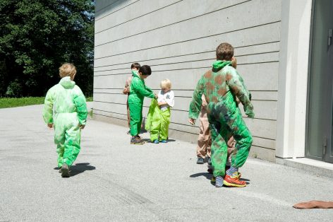 Several children wearing green and brown costumes walk and play near a modern concrete building on a sunny day. One child faces the wall, while others are moving around on the pavement. Trees and grass are visible in the background.