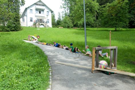 Children lay on a sloped path outside a white house, participating in an outdoor activity involving wooden frames and colorful balls on a lush green lawn surrounded by trees.