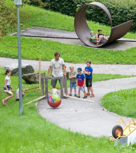 A group of children and an adult play an outdoor bowling game on a winding path, while three kids watch from inside a large metal sculpture in the background. Grass and trees surround the area.